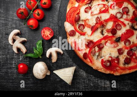 Pizza avec saucisses fumées, fromage, champignons, tomates cerises, poivrons et légumes verts sur une pierre et un tableau de craie noire, vue de dessus. Banque D'Images