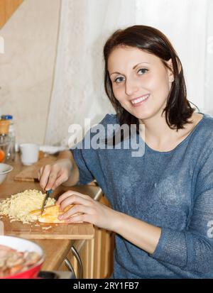 Jeune femme souriante coupant un fromage sur une planche à découper dans la cuisine Banque D'Images