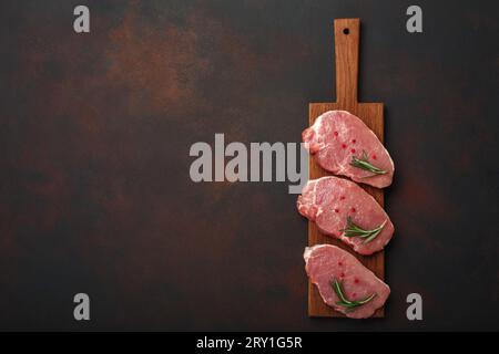 Morceaux de steak de porc cru sur la planche à découper avec romarin et poivron rouge sur fond brun rouillé. Vue de dessus avec espace pour votre texte. Banque D'Images