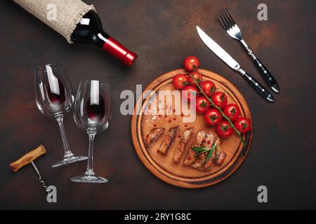 Tranches de steaks de porc grillés avec bouteille de vin, verre à vin, tire-bouchon, couteau, fourchette, pain noir, tomates cerises, ail, oignon et romarin sur rouillé Banque D'Images
