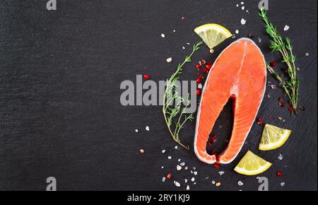 Steak de poisson de saumon cru frais avec des ingrédients de cuisine, des herbes et du citron sur fond noir, vue de dessus. Steak de saumon avec thym et citron sur table noire. Banque D'Images