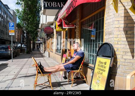 Un homme assis devant un magasin du coin, les pieds vers le haut, défilant sur un téléphone portable à Stockholm, en Suède. Photo : Rob Watkins Banque D'Images