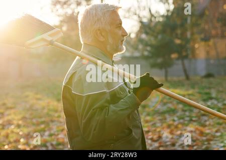 Travailleur de maintenance senior regardant loin, tout en gardant le balai sur l'épaule dans la chaude journée d'automne. Vue latérale de l'homme barbu en âge marchant avec balai, avec fond ensoleillé. Travaux d'entretien, concept d'automne. Banque D'Images