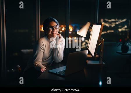 Heureuse femme d'affaires souriant à la caméra tout en travaillant tard dans son bureau. Femme professionnelle mettant dans de longues heures pour augmenter sa productivité et Banque D'Images