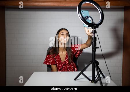 Souriante femme influenceuse commençant la vidéo en direct sur smartphone assis devant la lampe annulaire dans la pièce sombre Banque D'Images