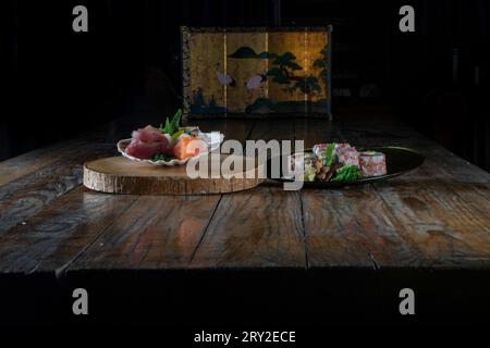 Sashimi appétissant de thon cru et de saumon dans une assiette blanche sur une tranche de bois tandis que des rouleaux de sushi faits avec du poisson avocat de riz dans une assiette noire et servi sur tabl Banque D'Images