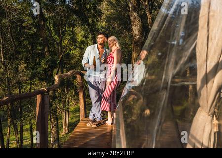 Corps entier de l'homme mûr et la femme clinking bouteilles de bière debout sur la terrasse en bois du dôme glamping dans la lumière du soleil Banque D'Images