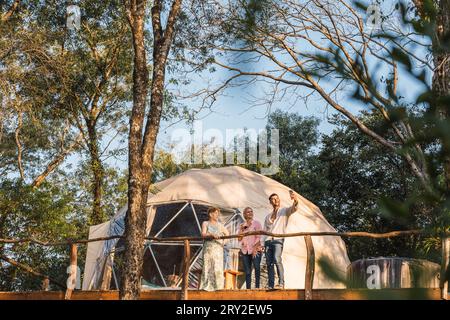 Jeune couple souriant avec femme mature regardant l'écran du téléphone portable et prenant selfie tout en se tenant ensemble près de tente de camp dans la forêt verte Banque D'Images