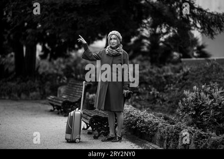 Une femme se tient sur la place avec une valise dans les mains et pointe son doigt quelque part. Photo en noir et blanc. Banque D'Images