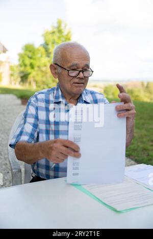 Un homme âgé est assis à un bureau dehors, travaillant avec des documents, remplissant ses formulaires médicaux, portant des lunettes, tenant un stylo. Banque D'Images
