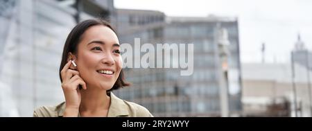 Prise de vue verticale d'une femme asiatique souriante dans un casque sans fil, aime écouter de la musique dans un casque, tient le téléphone portable Banque D'Images