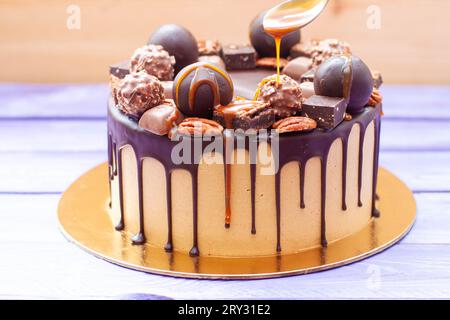 Gros plan de la main de femme versant de la sauce caramel salée sur le gâteau au chocolat, décoré de bonbons, truffes, biscuits et noix Banque D'Images