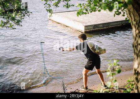 Vue arrière sur toute la longueur du garçon ramasser la corde du lac sur la rive Banque D'Images