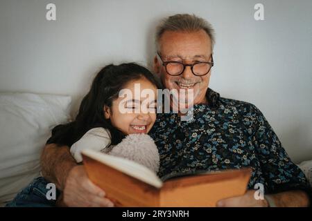 Homme âgé souriant lisant le livre de contes avec sa petite-fille sur le lit à la maison Banque D'Images