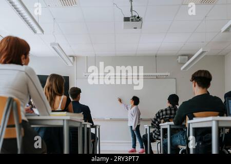 Étudiants assis à leur bureau pendant que l'enseignant écrit sur un tableau blanc en classe Banque D'Images