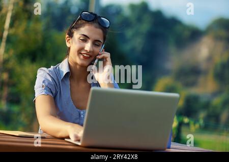 Travail à l'extérieur, jeune femme multitâches, téléphone en main, ordinateur portable ouvert. Ornée de blouse rayée bleue et blanche, des lunettes tiennent ses cheveux froncés. Arrière-plan Banque D'Images