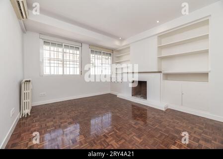 Salon dans l'appartement avec cheminée intégrée, barres métalliques sur les fenêtres, parquet rougeâtre brillant et étagères en bois blanc assorties Banque D'Images