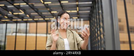 Une jeune fille asiatique se fait une joie de voir son appareil photo pour smartphone, dit bonjour à un ami lors d'un chat vidéo, appelant quelqu'un, debout dans la rue Banque D'Images