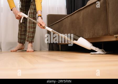 Méconnaissable, homme adulte pieds nus portant un pantalon à carreaux et un sol jaune à manches longues sous le canapé dans le salon. Homme avec la montre sur la main Cleanin Banque D'Images