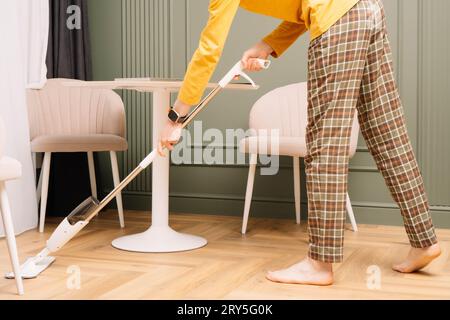 Humain méconnaissable en pantalon à carreaux et manches longues jaunes, regarder à la main laver le sol à l'aide d'une vadrouille près d'une table ronde à la maison. Femme de ménage à l'aise Banque D'Images