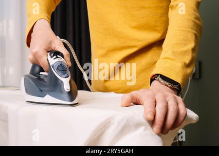 Gros plan de l'homme méconnaissable avec montre sur la main repasser chemise blanche sur pressboard à l'aide de flatiron électrique. Humain en manches longues jaune debout près Banque D'Images