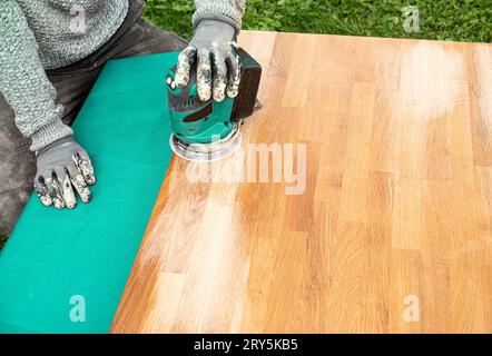 Vue rapprochée de la personne travaillant avec la ponceuse, ponçant les taches sur le vieux bloc de boucher endommagé sur la table en bois huilé. Concept de rénovation. Banque D'Images