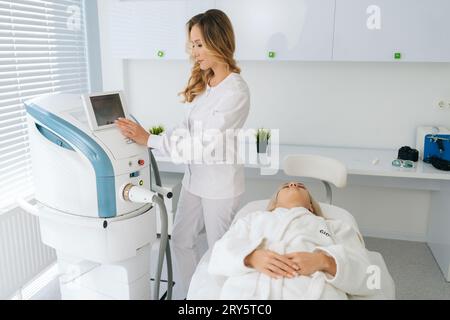 Portrait de spécialiste esthéticienne féminine ajuste l'appareil pour le photorajeunissement à l'aide de l'écran tactile. Femme cliente recevant le visage électrique stimulant Banque D'Images