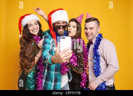 Groupe ou compagnie d'amis dans le chapeau de santa, chapeaux de fête, boas célébrant Noël, s'amuser, prendre des selfies et de vivre sur un fond jaune. Banque D'Images