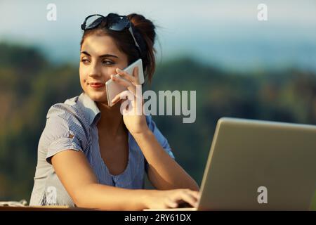 Dehors, jeune femme travaille énergiquement, conversant au téléphone, en utilisant un ordinateur portable. Porte un chemisier rayé bleu et blanc, des lunettes fixant ses cheveux, au milieu de la monture Banque D'Images