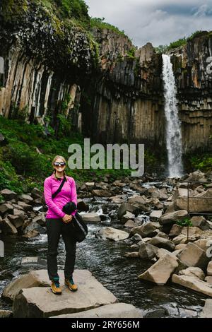 Femme sourit devant la cascade sur la falaise de basalte avec verdure Banque D'Images