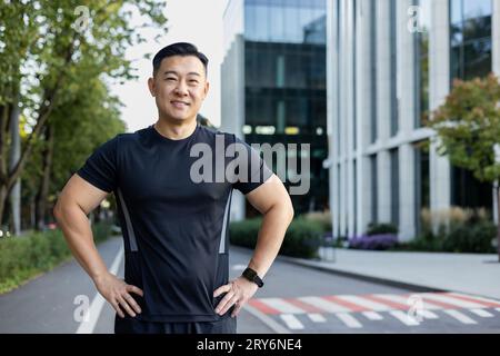 Portrait d'un jeune athlète asiatique musclé vêtu de vêtements de sport noirs debout dans une rue de la ville, mettant ses mains sur ses côtés et souriant à la caméra. Banque D'Images