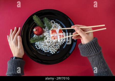 mains de femme tenant un cadeau avec des baguettes. Dans une assiette noire au lieu de la nourriture, il y a des perles blanches, une boule de Noël rouge et des branches vertes d'un arbre de Noël. Banque D'Images