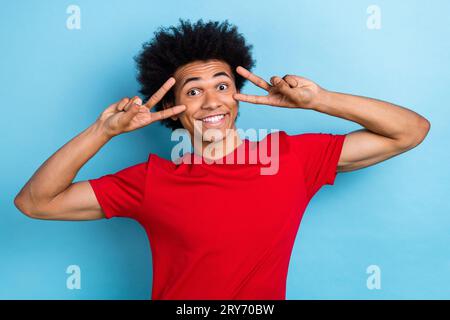 Portrait de drôle de cheveux bouclés jeune gars deux bras montrant des symboles de signe v près des yeux humeur flirty isolé fond de couleur bleue Banque D'Images
