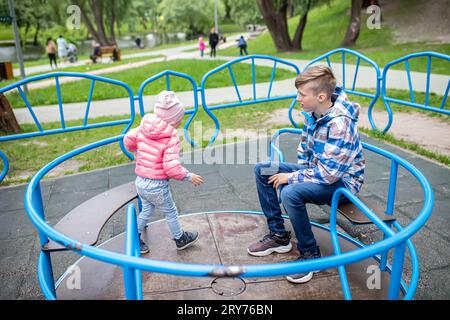 Frère et sœur montent un carrousel dans l'aire de jeux dans le parc de la ville. Les enfants s'amusent en été en plein air. Famille sur une promenade jouant ensemble Banque D'Images