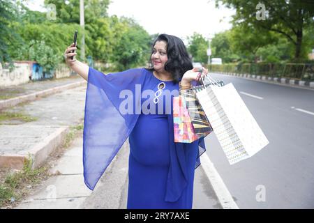 Jeune femme indienne de taille plus tenant des sacs à provisions prenant une photo selfie avec un téléphone intelligent à l'extérieur dans la rue. Femme en surpoids utilisant le téléphone. Banque D'Images