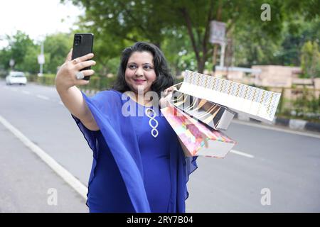 Jeune femme indienne de taille plus tenant des sacs à provisions prenant une photo selfie avec un téléphone intelligent à l'extérieur dans la rue. Femme en surpoids utilisant le téléphone. Banque D'Images