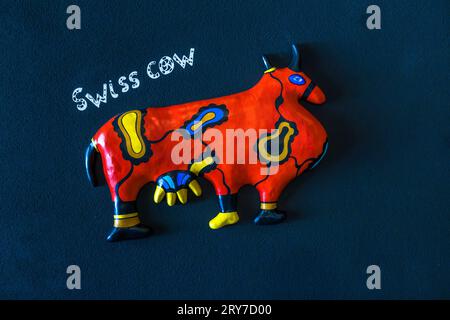 Une sculpture de vache plate et rouge-orange brillant portant l'étiquette manuscrite "Vache suisse" est accrochée à un mur sombre à l'Espace Jean Tinguely – Niki de Saint Phalle à Fribourg, Suisse. L'œuvre appartient à la série de reliefs "Rappel" (1997/98) de Niki de Saint Phalle. Installé depuis 1998 dans un ancien dépôt de tramway de Fribourg construit en 1900, le musée présente également l'œuvre majeure de Jean Tinguely, "Retable de l'Abondance occidentale et du Mercantilisme totalitaire" (1989/90). Banque D'Images