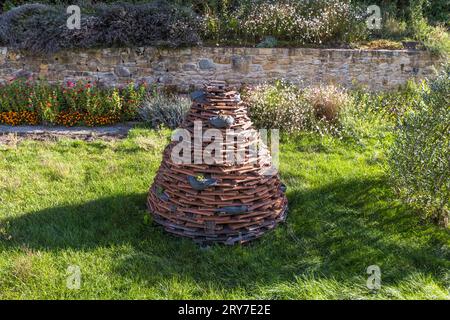 Une sculpture en forme de cône faite de tuiles empilées se dresse dans un jardin avec un parterre de fleurs et un mur de pierre sèche près de Morat (Murten), canton de Fribourg, Suisse. Banque D'Images
