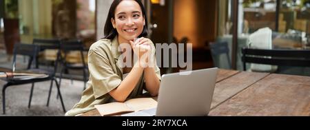 Portrait d'une fille asiatique travaille à l'extérieur dans un café, s'assoit avec un ordinateur portable, des études, sourires heureux Banque D'Images