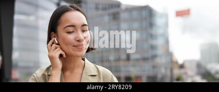 Prise de vue verticale d'une femme asiatique souriante dans un casque sans fil, aime écouter de la musique dans un casque, tient le téléphone portable Banque D'Images