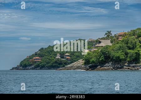 Zihuatanejo, Mexique - 18 juillet 2023 : demeures sur les falaises vertes boisées du rivage de l'océan montrant leurs toits rouges sous le paysage nuageux bleu Banque D'Images