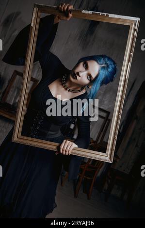 Portrait à l'intérieur de la magnifique fille goth en robe noire. Dame gothique aux cheveux bleus tient un cadre dans les mains Banque D'Images