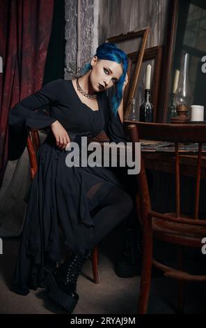 Portrait à l'intérieur de belle fille goth en robe noire. Dame gothique aux cheveux bleus assise sur un tabouret à la table. Look vintage Banque D'Images