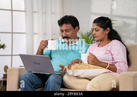 Couple indien détendu d'âge moyen utilisant un ordinateur portable tout en prenant un café à la maison - concept d'activité de loisirs, de connexion familiale et de convivialité Banque D'Images