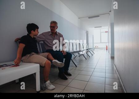 Dans un hôpital, un enfant attend avec son grand-père leur tour. Ils sont assis sur le banc et ont une conversation. Banque D'Images