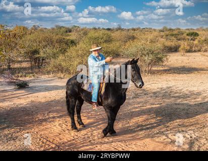 personne âgée avec une barbe et un chapeau de cow-boy habillé en denim, monté sur un cheval noir, menant le chemin, pointant vers les directions, sur un cheval safa Banque D'Images