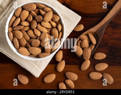 Un bol blanc rempli d’amandes biologiques conservées sur un tissu de coton blanc et d’amandes réparties autour d’une cuillère en bois Banque D'Images