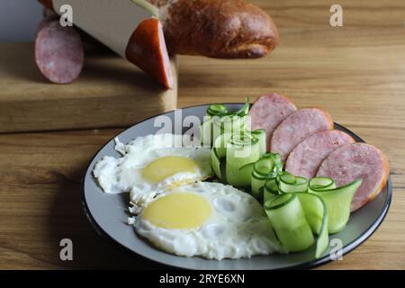 petit déjeuner traditionnel oeufs brouillés oeufs au plat deux saucisses au jambon et salade de concombre sur une assiette grise sur la table. a côté de lui se trouvent des produits pour la cuisson Banque D'Images