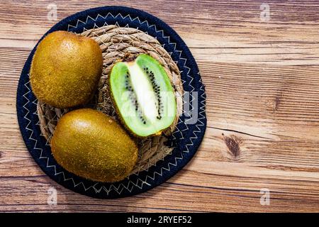 deux kiwis et un coupé en demi kiwi sur un tapis de paille circulaire à plat reposent sur un fond en bois isolé Banque D'Images