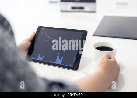 Femme regardant une tablette numérique avec graphique boursier et tenant une tasse de café dans une cuisine moderne. Commerce à domicile conce Banque D'Images